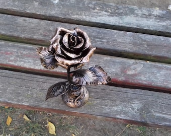 May include: A metal rose sculpture with a copper finish. The rose is in a vase and has leaves. The sculpture is on a wooden surface.
