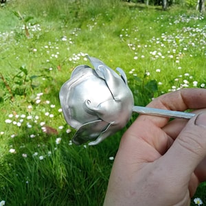 Pode incluir: Uma escultura de rosa em metal prateado, segurada em uma m&atilde;o. A rosa est&aacute; em um fundo de grama verde e pequenas flores brancas. A rosa de metal tem p&eacute;talas detalhadas e um caule.