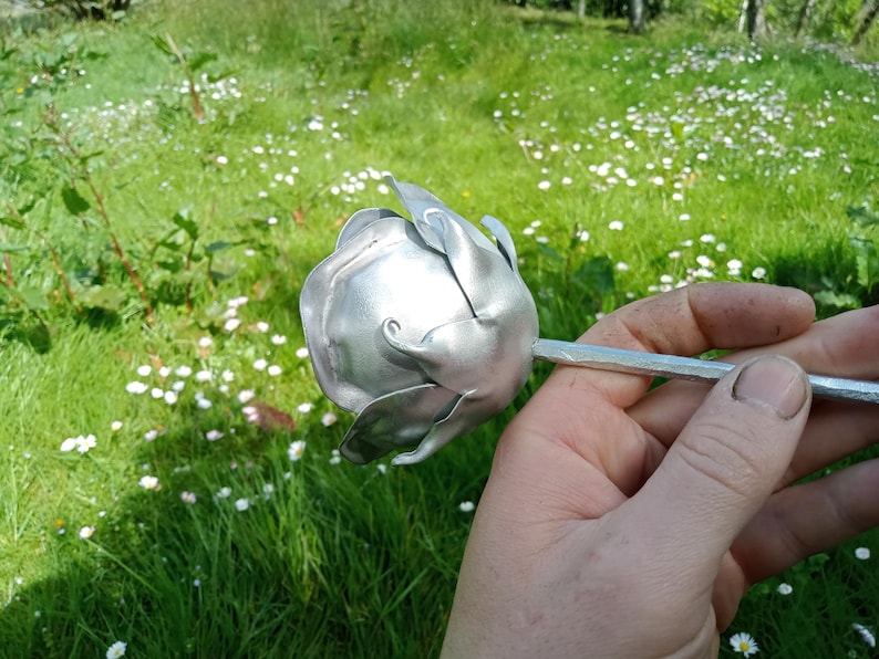 Pode incluir: Uma escultura de rosa em metal prateado, segurada em uma m&atilde;o. A rosa est&aacute; em um fundo de grama verde e pequenas flores brancas. A rosa de metal tem p&eacute;talas detalhadas e um caule.