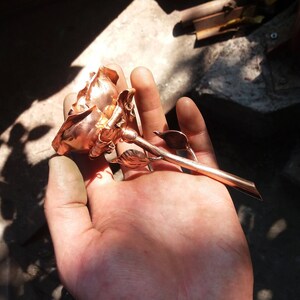 May include: A copper rose sculpture with a detailed stem and leaves. The rose is held in a hand, showcasing its intricate design.