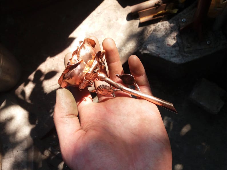 May include: A copper rose sculpture with a detailed stem and leaves. The rose is held in a hand, showcasing its intricate design.