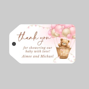 May include: Thank you for showering our baby with love! Aimee and Michael. A pink and white baby shower favor tag with a teddy bear in a hot air balloon.