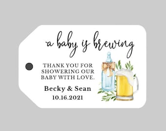 May include: A white tag with black text that reads "a baby is brewing". The tag also features a watercolor illustration of a baby bottle and a beer mug with a green leafy background. The text "Thank you for showering our baby with love." is also included, along with the names "Becky & Sean" and the date "10.16.2021".