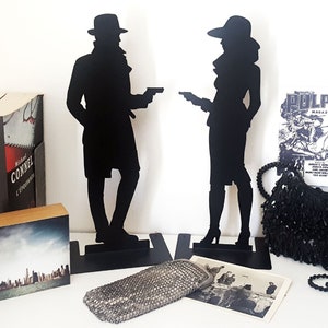 Op de afbeelding: Twee zwarte metalen boekenleggers in de vorm van silhouetten van een man en een vrouw in vintage stijl. De man draagt een fedora en een trenchcoat en houdt een pistool vast. De vrouw draagt een hoed en een jurk en houdt een pistool vast. De boekenleggers staan op een wit oppervlak met een zwarte handtas en een tijdschrift met het woord "PULP" op de omslag.