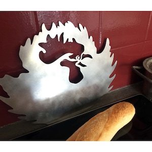 Może przedstawiać: Srebrna metalowa sylwetka koguta z ozdobnym wzorem. Koguć jest zamontowany na ścianie nad czarnym blatem.