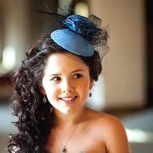 May include: A young woman with long, dark curly hair wearing a blue and black dress with a blue hat and a necklace with a heart pendant.