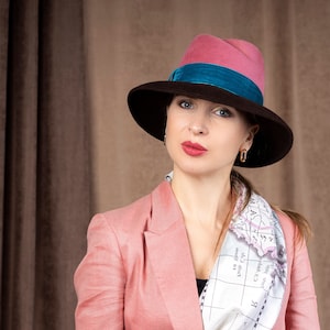 May include: A woman in a pink blazer, a map-print scarf, and a fedora hat. The hat is brown with a pink crown and a teal band. The background is a brown curtain.