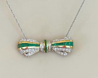 Puede incluir: Un collar con colgante en forma de lazo que presenta una banda central de esmeraldas verdes engastadas en canal, flanqueada por filas de diamantes blancos en pavé, todo montado en oro amarillo. El colgante cuelga de una fina cadena de metal plateado.
