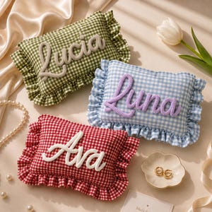 May include: Three rectangular cushions with ruffled edges and a gingham pattern in olive green, light blue, and red. Each cushion features a name written in raised, knitted script lettering: Lucia in beige, Luna in lavender, and Ava in white.