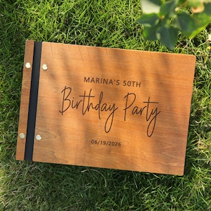 May include: A wooden sign with the text "MARINA'S 50TH Birthday Party 06/19/2026" engraved in a stylish font. The sign is rectangular, with a dark black spine and gold-colored rivets. The sign is lying on a bed of green grass.