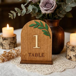May include: Wooden table number one with a green leaf accent. The number and the word "TABLE" are in a lighter wood tone against the darker wood background. The table number is displayed on a white lace tablecloth.