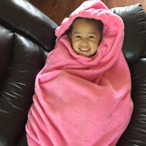 Op de afbeelding: Een kind gewikkeld in een zachte, roze deken met capuchon, lacht naar de camera. De deken is zacht en bedekt het lichaam van het kind, wat een gezellige en comfortabele uitstraling creëert. De achtergrond is een donkerbruine leren bank.