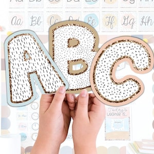 May include: The letters A, B, and C are held in front of a classroom display. The letters have a white background with black dashes and a gold glitter border. The letter A has a light blue border, while B and C have a tan border. The background includes other letters and a weather chart.