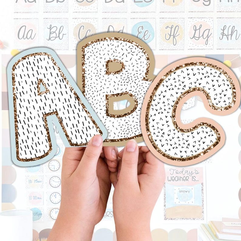 May include: The letters A, B, and C are held in front of a classroom display. The letters have a white background with black dashes and a gold glitter border. The letter A has a light blue border, while B and C have a tan border. The background includes other letters and a weather chart.