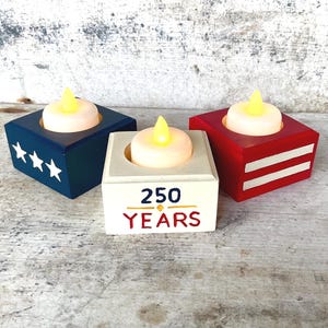 May include: Three wooden candle holders in patriotic colors. One is navy blue with white stars, another is red with white stripes, and the third is white with the text "250 YEARS". Each holds a battery-operated candle with a yellow flame.