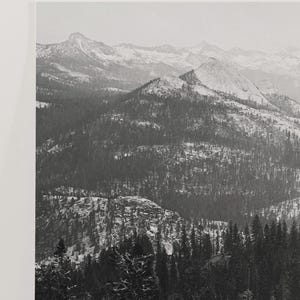 May include: A black and white photograph of a snow-covered mountain range with a dense forest in the foreground.