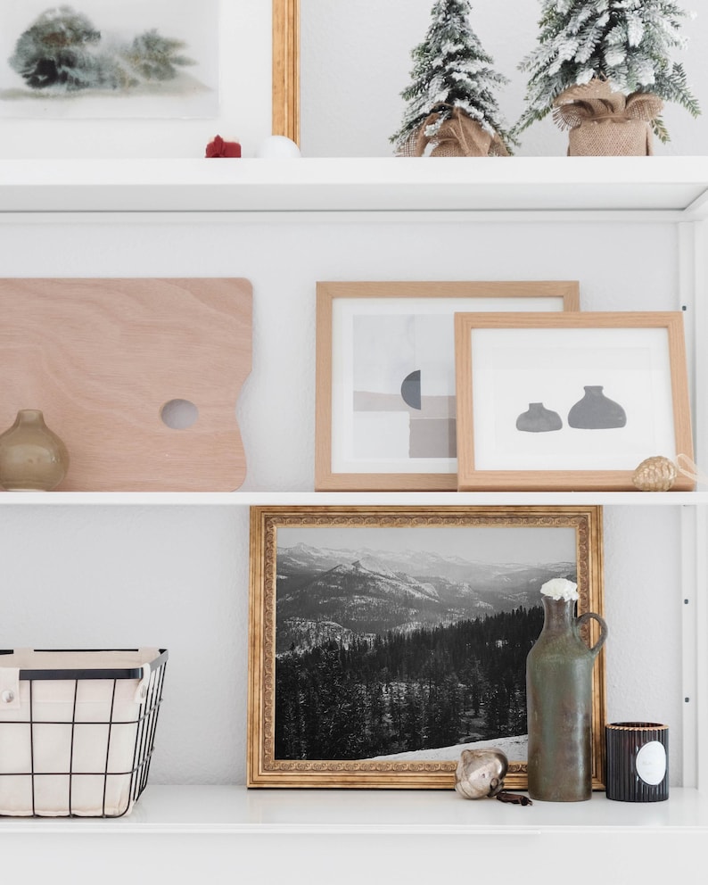 May include: A white shelf with a variety of items, including a wooden cutting board, framed artwork, a black and white photo of a mountain range, a ceramic vase, a glass bottle, and a small decorative object. The shelf is decorated with two small artificial Christmas trees.