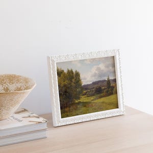 Op de afbeelding: Een wit fotolijstje met een landschapschilderij van een groen veld met bomen en een bewolkte lucht.