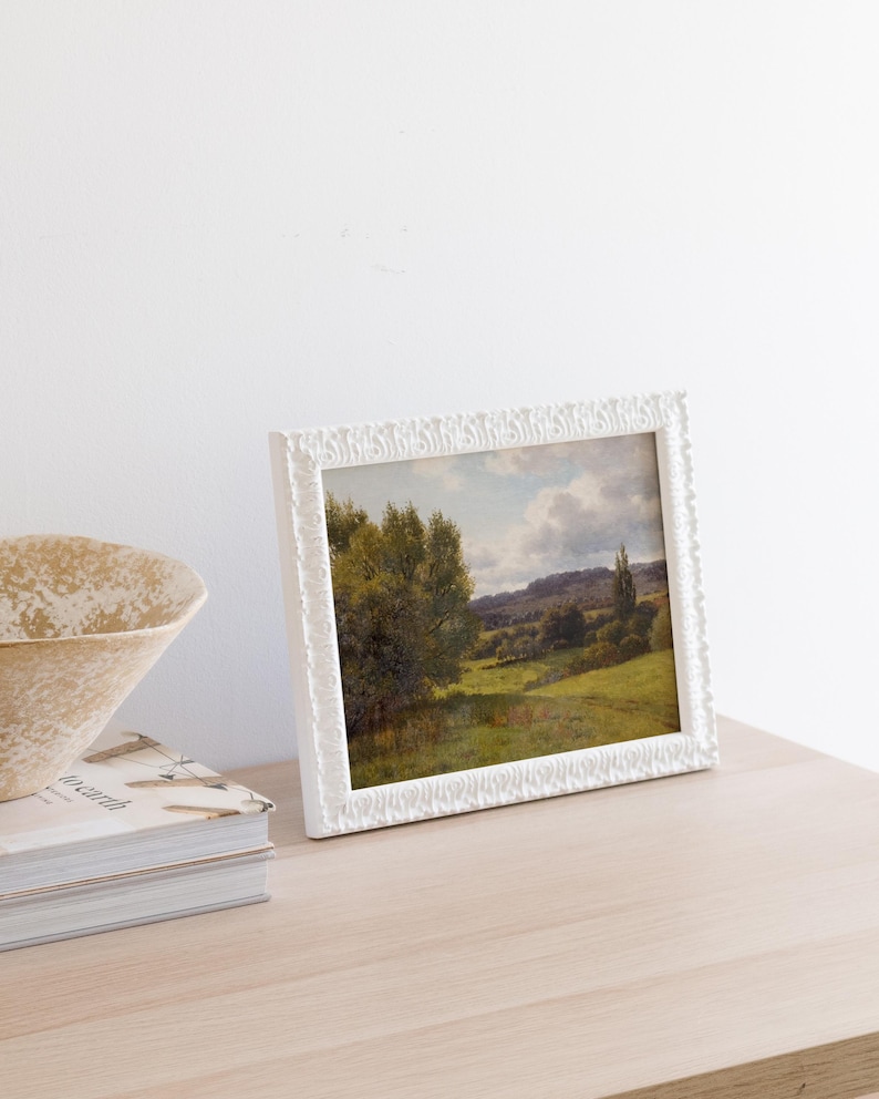 Op de afbeelding: Een wit fotolijstje met een landschapschilderij van een groen veld met bomen en een bewolkte lucht.