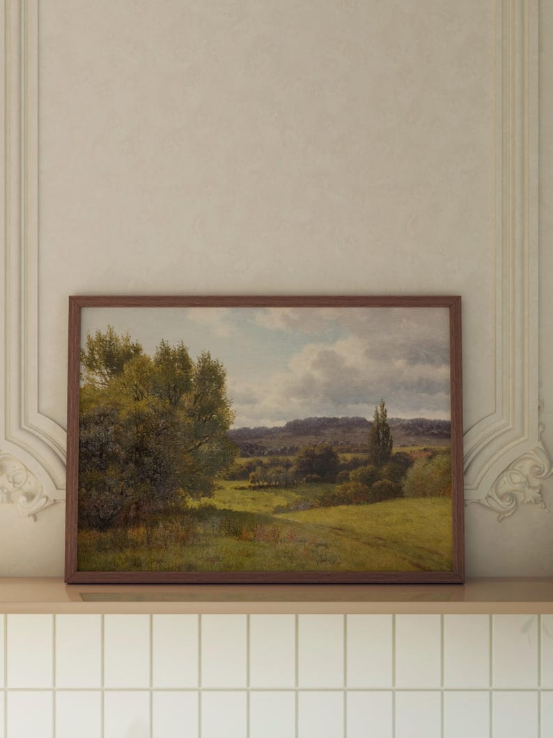 Op de afbeelding: Een ingelijst landschapsschilderij dat een landelijk tafereel voorstelt met een weelderig groen veld, bomen en een heuvel in de verte onder een bewolkte hemel.