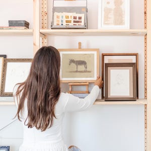 Puede incluir: Una mujer se encuentra frente a una estanter&iacute;a de madera con obras de arte enmarcadas. Las obras de arte incluyen un dibujo de un burro, un dibujo de una persona y un dibujo de dos perros. La estanter&iacute;a est&aacute; llena de otras obras de arte enmarcadas y una bandeja de metal con muestras de pintura.