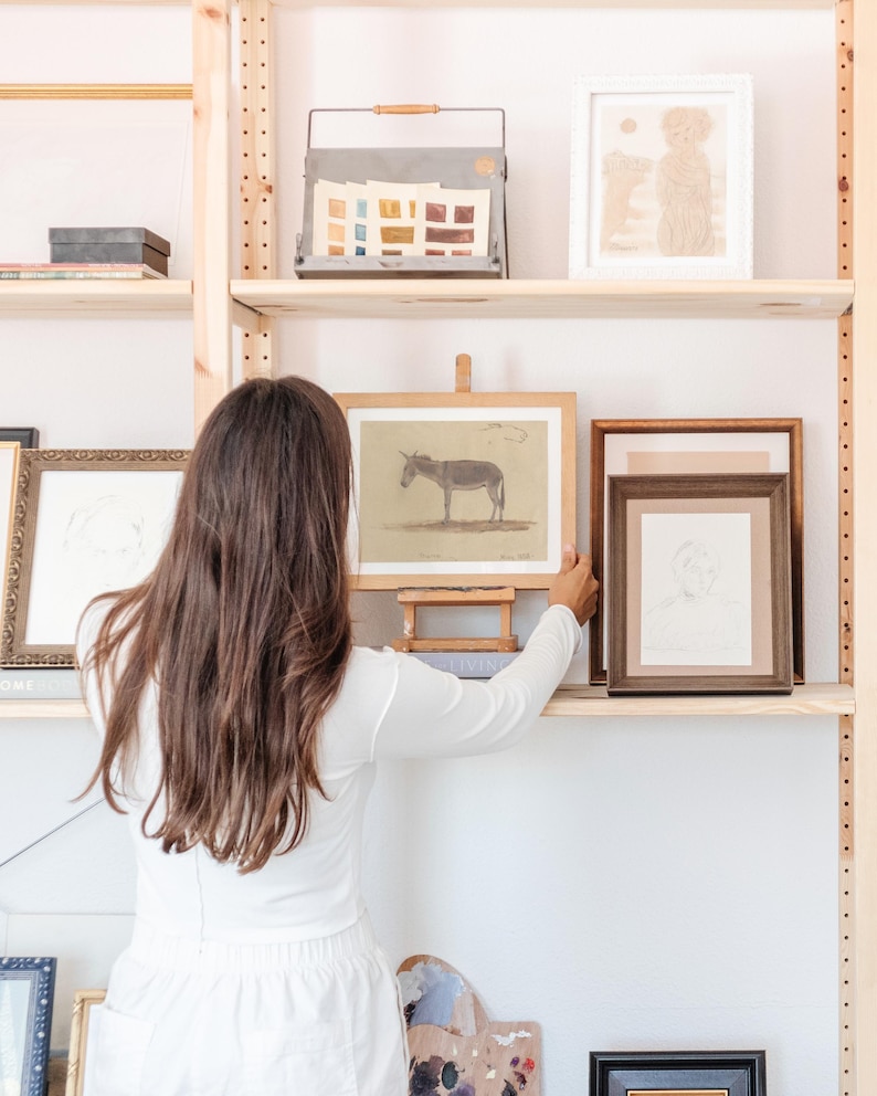 Puede incluir: Una mujer se encuentra frente a una estanter&iacute;a de madera con obras de arte enmarcadas. Las obras de arte incluyen un dibujo de un burro, un dibujo de una persona y un dibujo de dos perros. La estanter&iacute;a est&aacute; llena de otras obras de arte enmarcadas y una bandeja de metal con muestras de pintura.