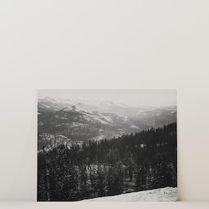 May include: A black and white photograph of a snowy mountain range with a forest in the foreground.
