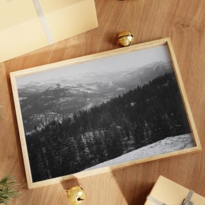 May include: A framed black and white photograph of a snowy mountain range with a forest in the foreground. The frame is made of light wood and is sitting on a wooden surface.