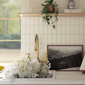 May include: A gold faucet with two handles is mounted on a white tiled countertop. A white ceramic sink is partially filled with white flowers. A framed black and white photograph of a mountain range is on the countertop. The text "The  Maconde Book of JAPAN at 100" is on a shelf above the countertop.