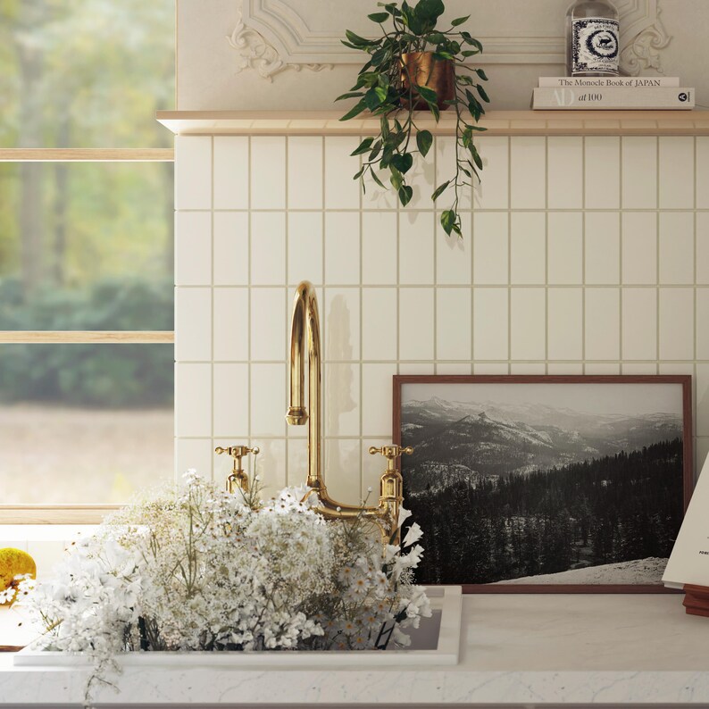 May include: A gold faucet with two handles is mounted on a white tiled countertop. A white ceramic sink is partially filled with white flowers. A framed black and white photograph of a mountain range is on the countertop. The text "The  Maconde Book of JAPAN at 100" is on a shelf above the countertop.