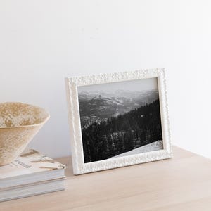 May include: A white picture frame with a black and white photograph of a mountain range. The frame has a decorative, textured border.