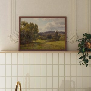 Op de afbeelding: Een ingelijst landschapschilderij van een groen veld met bomen en een bewolkte lucht. Het schilderij hangt aan een witte betegelde muur.
