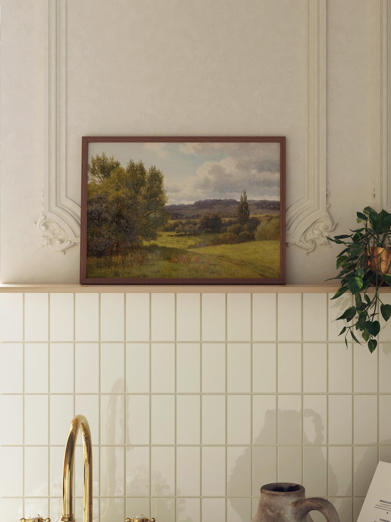 Op de afbeelding: Een ingelijst landschapschilderij van een groen veld met bomen en een bewolkte lucht. Het schilderij hangt aan een witte betegelde muur.