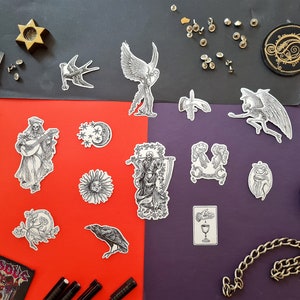 Könnte beinhalten: Eine Sammlung von schwarz-weißen Aufklebern mit verschiedenen Illustrationen, darunter ein Rabe, ein Skelett, das Laute spielt, eine Sonne, ein Mond und eine Hand, die einen Kelch hält. Die Aufkleber sind auf einem roten und violetten Hintergrund angeordnet.