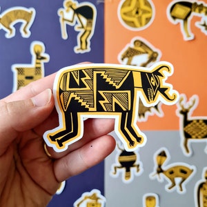 May include: A black and yellow patterned sticker of a stylized animal with a long snout and horns. The sticker is on a white background and is being held by a hand.