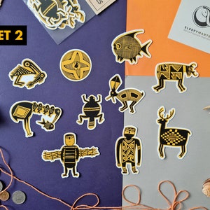 May include: Set of 10 black and gold patterned stickers featuring various animal and geometric designs. The stickers are on a purple and gray background with a small piece of burlap fabric and some seashells.