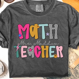 May include: Dark gray t-shirt with the words "MATH Mrs. Maddow TEACHER" in colorful, playful lettering. The design includes stars, a lightning bolt, and a smiley face. The shirt is a comfortable, casual style.