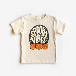 May include: A cream coloured t-shirt with a black circle graphic that says "Spooky Vibes" with three orange jack-o'-lantern faces below the text.