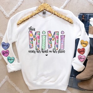 May include: White sweatshirt with the text "this MIMI wears her heart on her sleeve" printed on the front. The sleeves have colorful heart-shaped patches with names on them.