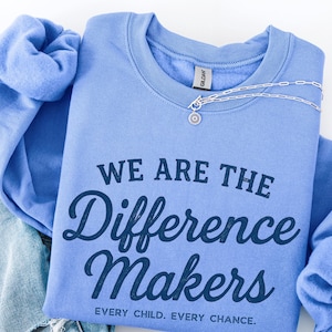 May include: A light blue sweatshirt with the text "WE ARE THE Difference Makers" in dark blue. The sweatshirt has long sleeves and a crew neck. A silver necklace with a small pendant is draped across the top. Denim jeans are visible on the left.