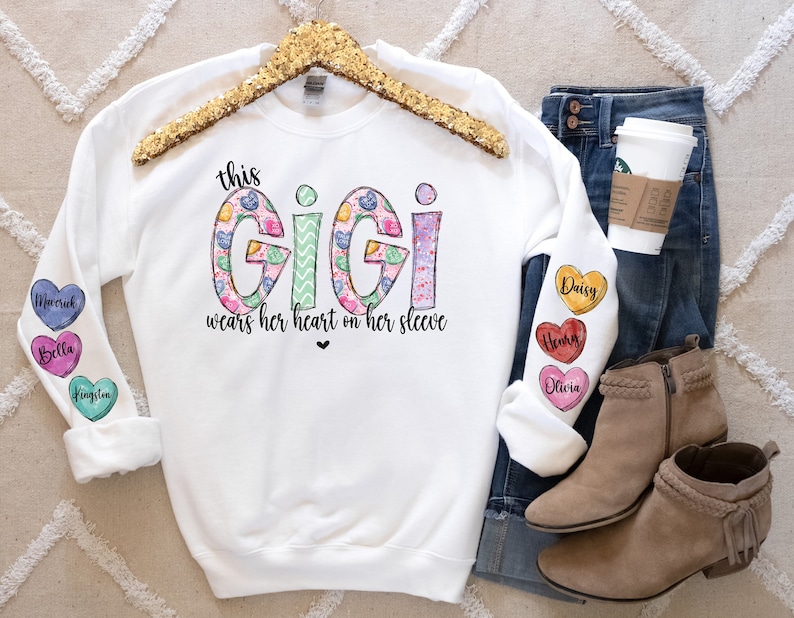 May include: White sweatshirt with the text "this GiGi wears her heart on her sleeve" printed on the front. The word "GiGi" is decorated with a floral pattern. The sleeves have colorful heart-shaped patches with names printed on them.