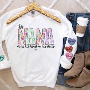 May include: White sweatshirt with the text "this NANA wears her heart on her sleeve" in pink, green, and yellow. The sleeves have colorful heart-shaped patches with names on them, including Daisy, Bella, Kingston, Mecerick, Henry, and Olivia.