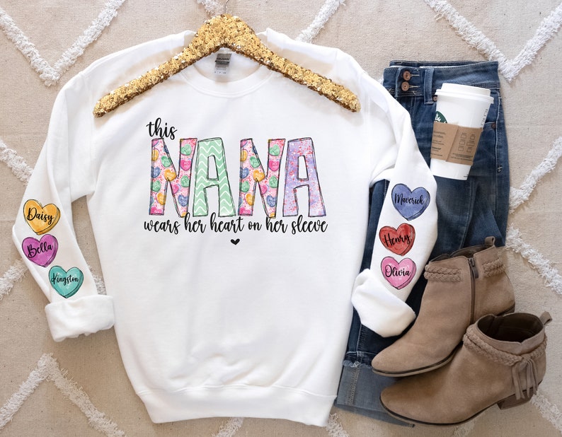 May include: White sweatshirt with the text "this NANA wears her heart on her sleeve" in pink, green, and yellow. The sleeves have colorful heart-shaped patches with names on them, including Daisy, Bella, Kingston, Mecerick, Henry, and Olivia.