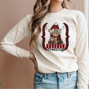 May include: A long-sleeved, cream-colored t-shirt with a cartoon highland cow wearing a red and black plaid scarf and a red hat. The cow is holding a mug of hot chocolate with marshmallows. The text "Udderly freezing" is printed below the cow.
