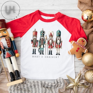 May include: A white and red baseball tee shirt with a graphic of four nutcracker soldiers and the text "What's Crackin?"