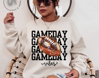 May include: A cream colored sweatshirt with the text "GAMEDAY" repeated four times with a glittery brown football in the center. The text "vibes" is written below the football.