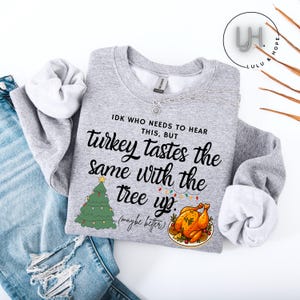 May include: A light gray sweatshirt with the text "IDK WHO NEEDS TO HEAR THIS, BUT turkey tastes the same with the tree up. (maybe better)". The design includes a Christmas tree and a roasted turkey.