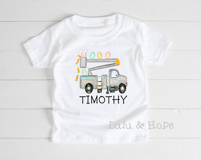 K&ouml;nnte beinhalten: Wei&szlig;es T-Shirt mit einem grauen LKW, der Ostereier und Karotten transportiert. Der LKW hat einen Ausleger und Hasenohren oben drauf. Der Text "TIMOTHY" ist unter dem LKW gedruckt.