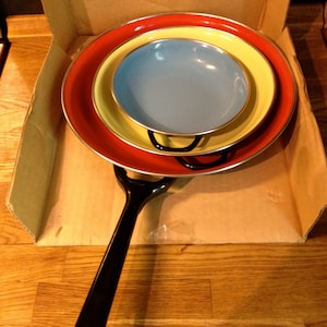 Puede incluir: Un juego de vajilla de esmalte colorido en una caja de cart&oacute;n. El juego incluye un plato rojo, un plato amarillo y un cuenco azul, todos con bordes plateados. Un asa negra est&aacute; unida al plato inferior. Los platos est&aacute;n anidados.