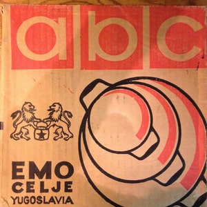 Puede incluir: Una caja de cart&oacute;n vintage con las letras "ABC" en un rect&aacute;ngulo rojo en la parte superior. Debajo, la caja presenta un gr&aacute;fico de una olla con detalles en rojo y el texto "EMO CELJE YUGOSLAVIA". La caja tiene un escudo de le&oacute;n.